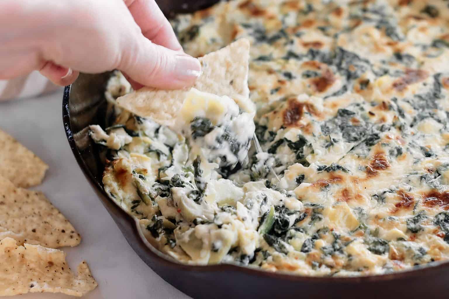 A hand dips a tortilla chip into a skillet filled with hot, creamy spinach and artichoke dip, featuring melted cheese on top. A few tortilla chips are positioned around the skillet on a white surface.
